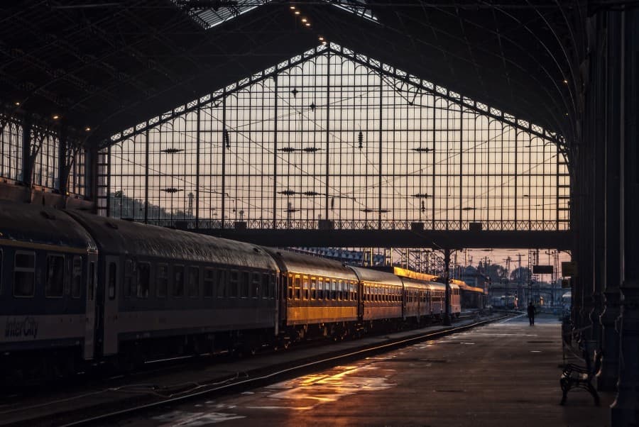 Die hohen Glasfenster des Bahnhofs in Budapest leuchten im Sonnenuntergang. Zugwagen stehen auf den Gleisen, Passagier wartet rechts. Perfekter Start für einen entspannten Urlaub in Budapest.