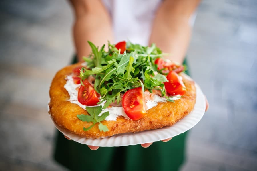 Ein goldbrauner Langos mit Sauerrahm, Rucola und halbierten Kirschtomaten auf einem weißen Pappteller gehalten von zwei Händen. Der Hintergrund ist verschwommen und zeigt einen grauen Boden.
