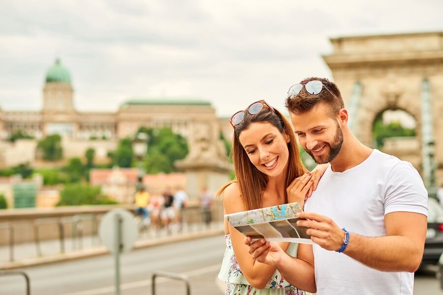 Ein lächelndes Paar, das eine Karte betrachtet, steht auf einer Brücke in Budapest. Im Hintergrund sind die grüne Kuppel und die imposante Architektur des Budapester Budaer Schlosses zu sehen. Perfekt für Urlaub in Budapest.