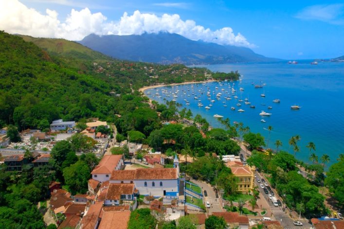 Luftaufnahme von Ilhabela, Brasilien, mit grünen Hügeln im Hintergrund, zahlreichen Segelbooten auf blauem Wasser und einer kleinen Küstenstadt.
