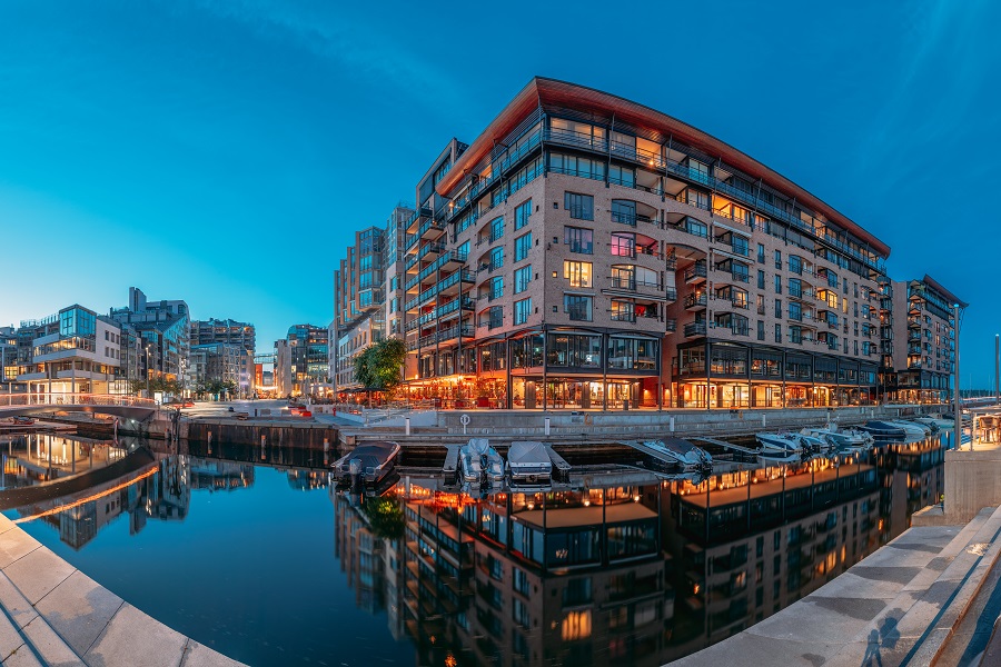 Ein Gebäude im Hafenviertel Aker Brygge mit einem Fischaugenobjektiv fotografiert. Es ist bereits dunkler, aber noch nicht Nacht. In dem Gebäude brennt Licht und es spiegel sich im Wasser davor.