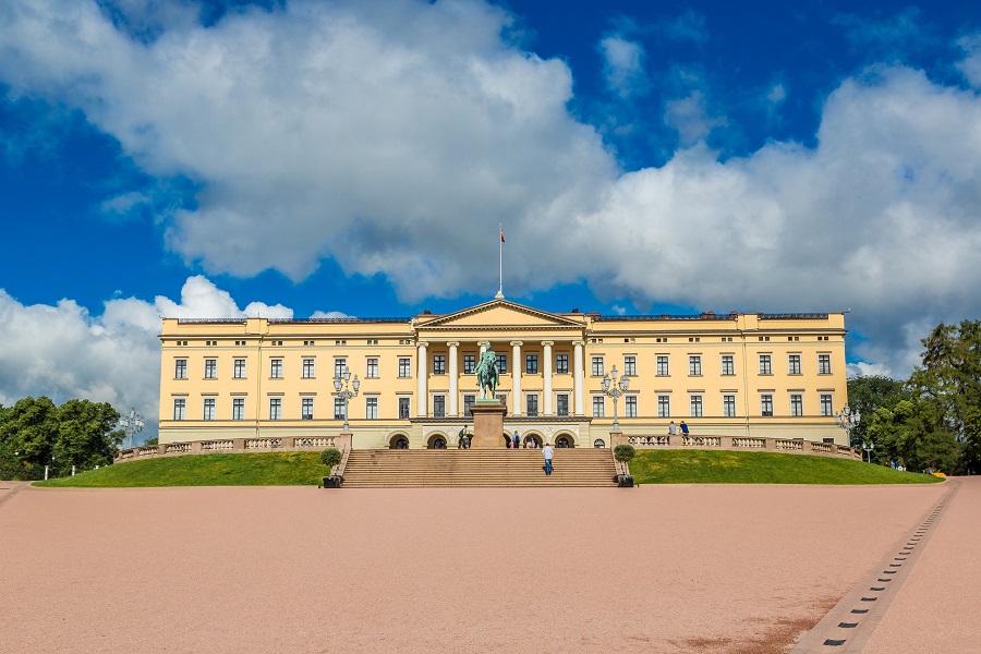 Foto des königlichen Schlosses von Oslo. Der Platz im Vordergrund ist leer und aus braun-rotem Stein. Treppen führen zu einem prunvollen, beigen Gebäude hinauf. Vor dem Gebäude steht eine Statue. Der Himmel ist blau mit wenigen Wolken.