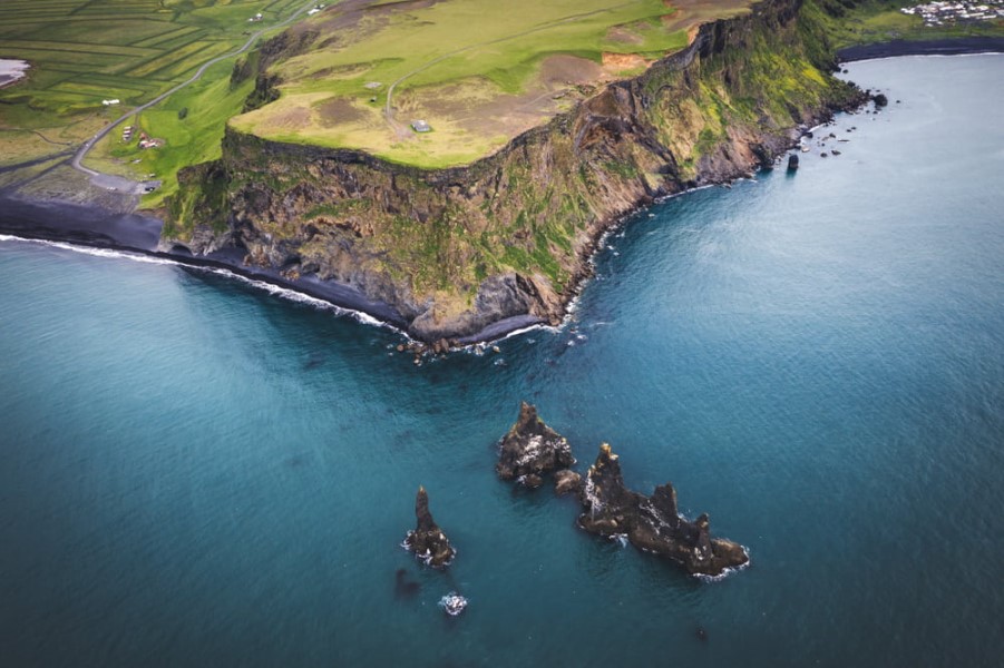 Foto einer Klippe in Island. Im unteren Teil des Bildes ist das Meer mit ein paar Felsen darin, in der oberen Hälfte sieht man die grün bewachsene Klippe.