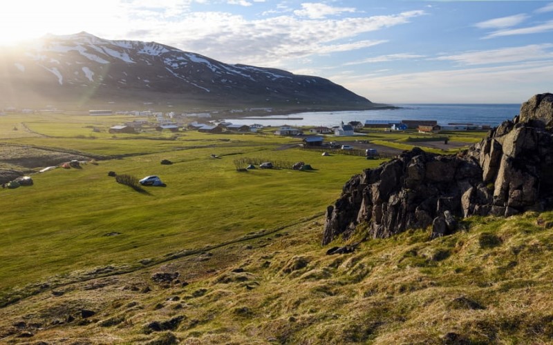 Foto des Dorfes Bakkagerdi in Island. Man sieht sehr viel Landschaft und nur wenige Häuser. Auf der rechten Seite des Bildes ist ein Teil des Meeres zu sehen. Im Hintergrund ist ein hoher Berg mit ein wenig Schnee.