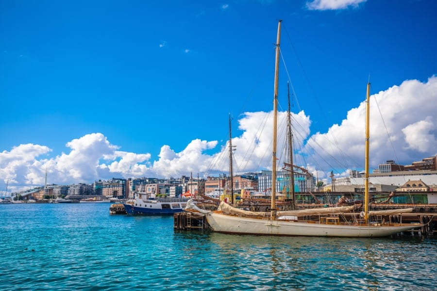 Bild vom Osloer Hafen vom Wasser aus. Im Vordergrund ist ein Segelboot zu sehen, dahinter sind noch mehr zu erahnen. Im Hintergrund ist die erste bunte Häuserreihe des Hafenviertels Aker Brygge zu erkennen.