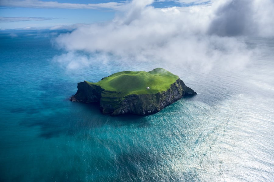 Luftaufnahme der Insel Heimaey in Island. Sie ragt aus dem türkisblauen Wasser. Die Klippen sind steinig, die Fläche der Insel ist saftig grün. Eine Wolke zieht über die Insel.