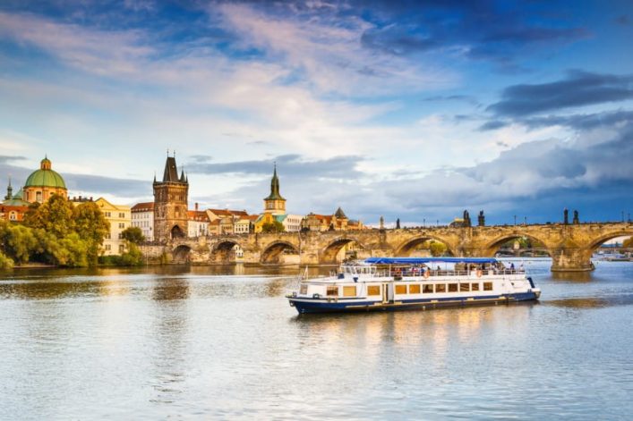 Städtische Skyline mit historischen Gebäuden rechts und links einer steinernen Brücke. Im Vordergrund fährt ein Flusskreuzfahrtschiff vorbei. Perfekt für Oktober Kreuzfahrten bei herbstlicher Stimmung.