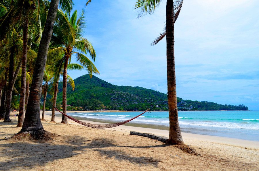 Palmen säumen einen sonnigen Sandstrand, an dem eine Hängematte befestigt ist. Im Hintergrund liegt ein grüner Hügel, das ruhige Meer glitzert darunter – ideal für einen Familienurlaub in Thailand.