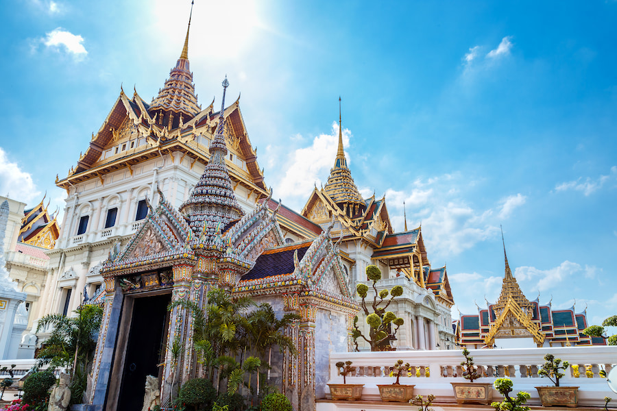 Der prächtige Königspalast in Thailand leuchtet in der Sonne mit kunstvollen Dächer und goldenen Details, ideal für einen Familienurlaub in Thailand. Im Vordergrund sind bunte Verzierungen und Pflanzen zu sehen.