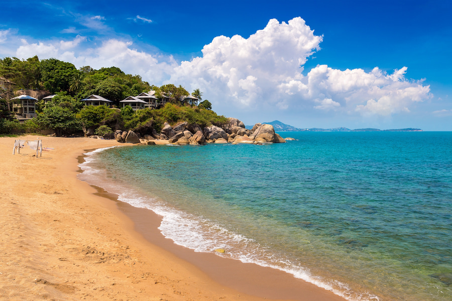 Die Coral Cove Beach auf Koh Samui in Thailand mit klarem, türkisblauem Wasser, goldenem Sandstrand und felsigen Vorsprüngen. Im Hintergrund sind grüne Hügel mit Villen zu sehen. Ideal für einen Familienurlaub.