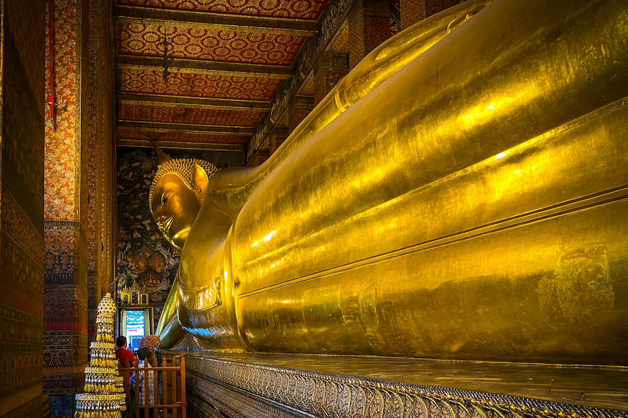 Goldener, liegender Buddha im Wat Pho Tempel in Bangkok, Thailand, ein beliebtes Ziel für Familienurlaub in Thailand. Die Statue nimmt den größten Teil des Bildes ein, links daneben stehen bewundernde Besucher.