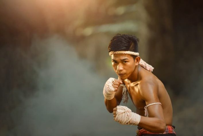 Muay-Thai-Kämpfer in Angriffshaltung, mittig im Bild, mit Bandagen an Händen und Stirn. Sonnenlicht fällt von links ein, Hintergrund ist verschwommen und rauchig, was eine intensive Atmosphäre schafft.
