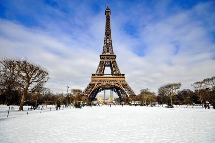 Schneebedeckter Rasen im Vordergrund, der Eiffelturm ragt imposant in der Mitte empor. Kahle Bäume säumen den Weg, vereinzelt spazieren Menschen. Der Himmel ist bewölkt mit blauen Lücken.