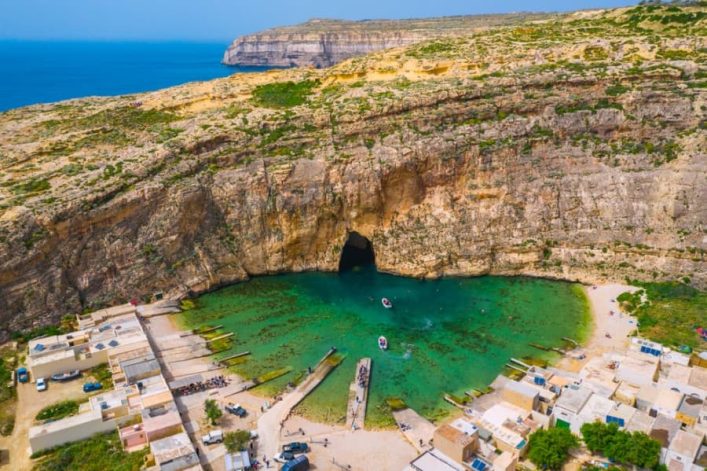 Steile Felsklippen umgeben die türkisfarbene Dwejra-Bucht auf Gozo. Im Vordergrund kleine Gebäude und Boote an Land. In der Mitte führt ein Höhleneingang ins Meer. Klare, sonnige Atmosphäre.