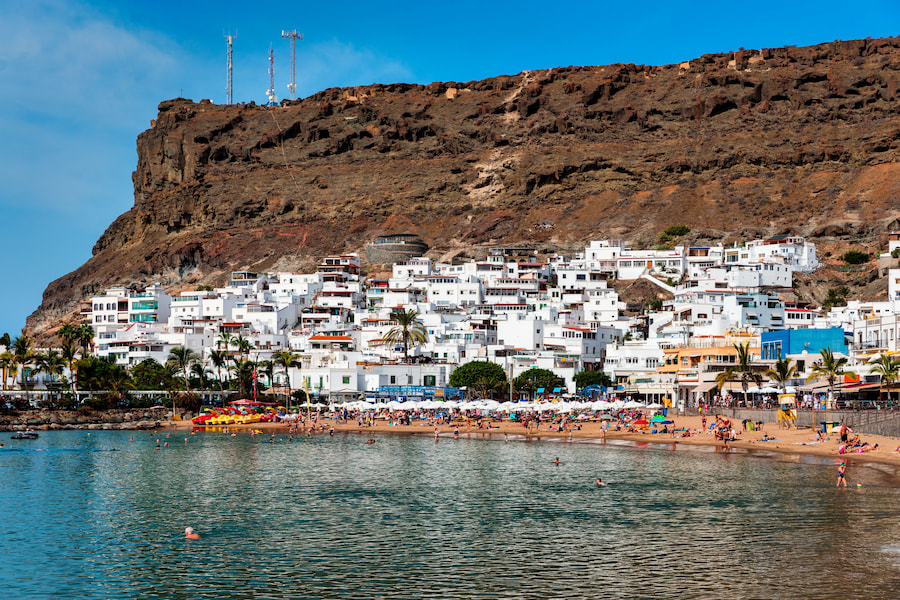 Heller Sandstrand mit Familien, umrahmt von bunten Häusern am Hang und Palmen. Klippen im Hintergrund. Perfekt für Familienurlaub auf Gran Canaria. Ruhiges Wasser im Vordergrund.